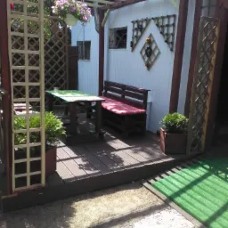 Zadaszony taras z drewnianą podłogą, stołem z zielonym blatem i ławką z czerwonym siedziskiem, dekoracje z kratki na ścianie domku i wiszące kwiaty w doniczce.