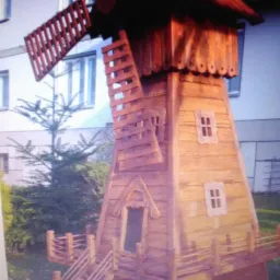 Ozdobny, drewniany model wiatraka z miniaturowymi schodami i okienkami, stojący na trawie przed budynkiem mieszkalnym.