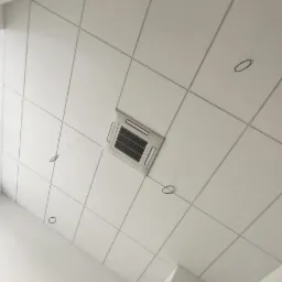 Biały sufit kasetonowy z wbudowaną kratką wentylacyjną, widoczne otwory na oświetlenie i fragment okna z parapetem.