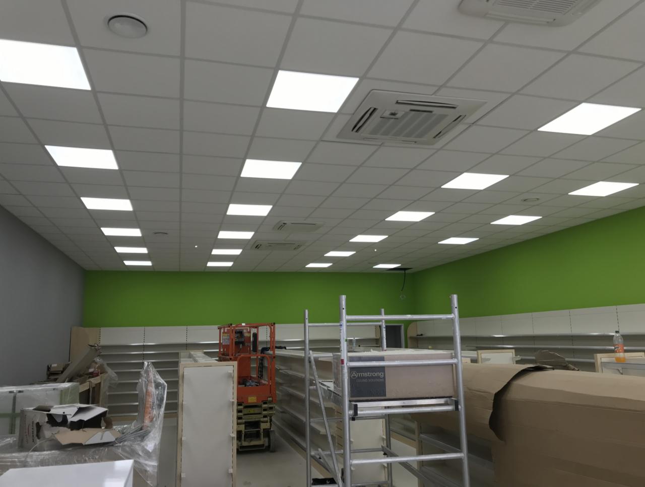 Wnętrze komercyjne w trakcie prac wykończeniowych: sufit podwieszany z kwadratowymi panelami oświetleniowymi, świeżo pomalowane ściany w odcieniach szarości i soczystej zieleni, regały sklepowe...