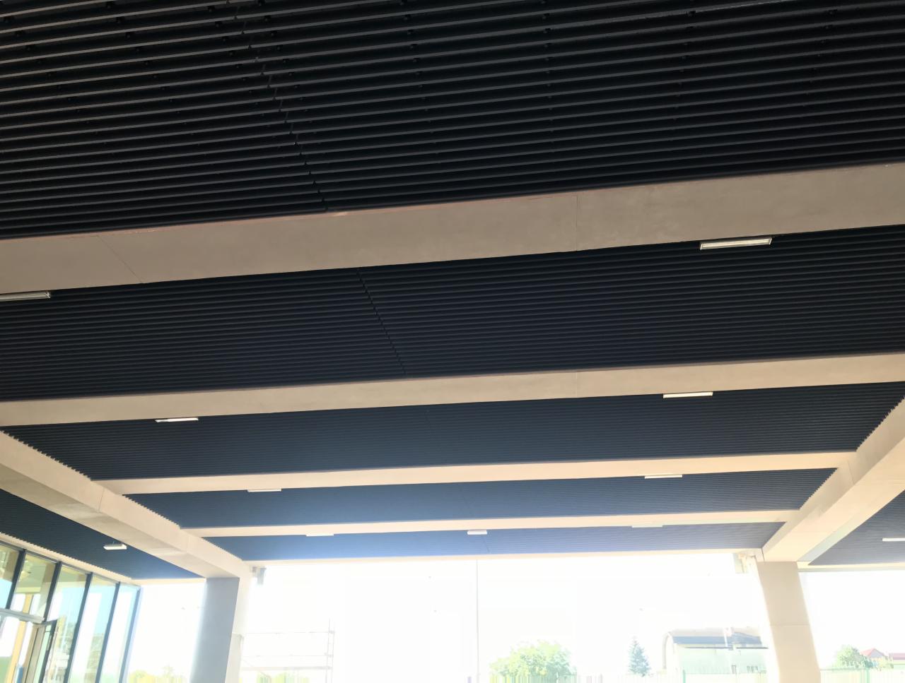 Czarne, żłobkowane panele sufitowe zamontowane w biurze z widocznymi białymi belkami konstrukcyjnymi i oświetleniem LED.