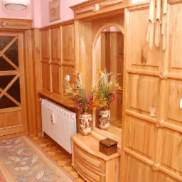 Drewniana zabudowa przedpokoju z szafą, lustrem, komodą i drzwiami, w ciepłym odcieniu drewna, dekorowana kwiatami w wazonie.