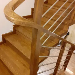 Drewniane schody z jasnego drewna z metalowymi poręczami, widok z góry, z zaokrąglonym elementem poręczy na pierwszym planie.