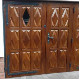 Dwuskrzydłowe, zdobione drzwi wejściowe z drewna w ciepłym odcieniu brązu, z romboidalnymi wzorami i małymi okienkami, otoczone czarnymi, kutymi elementami dekoracyjnymi.