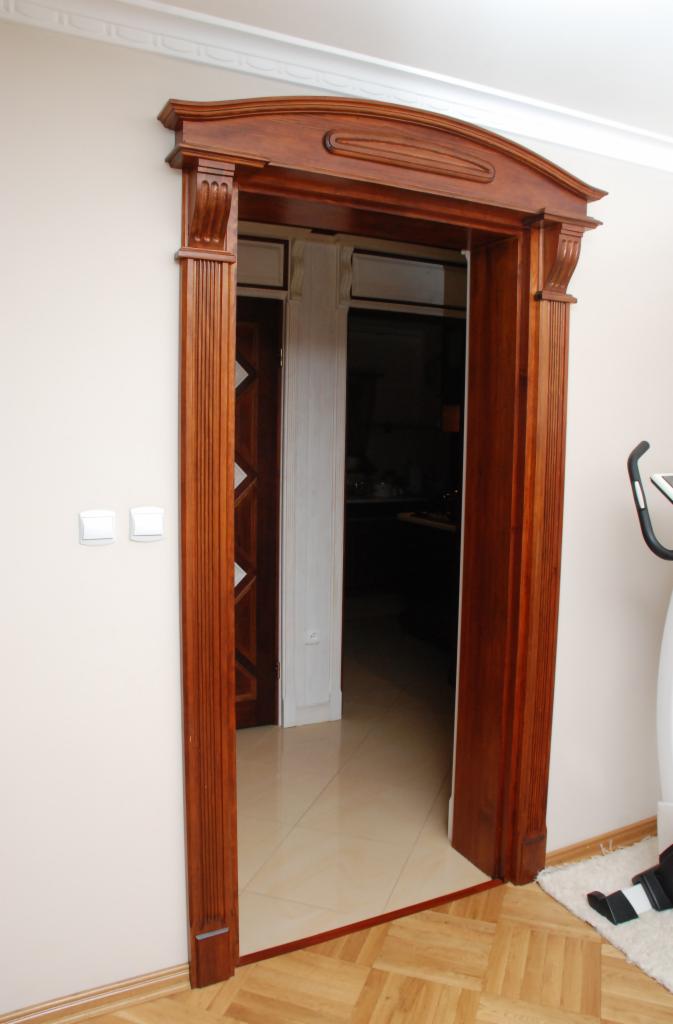 Rama drzwiowa z ciemnego drewna z dekoracyjnymi elementami, widok na sąsiednie pomieszczenie z drzwiami i jasnymi płytkami podłogowymi.