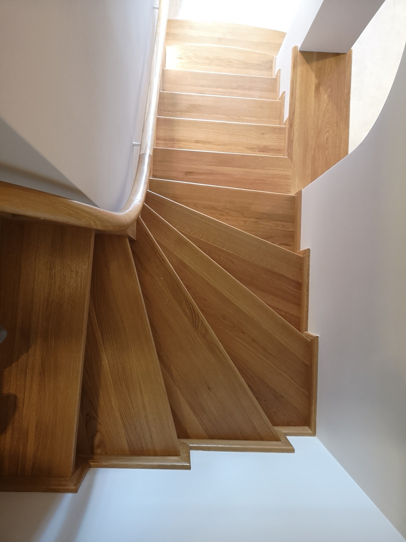 Widok z góry na drewniane schody zabiegowe z jasnego drewna, z delikatnym oświetleniem z góry i zaokrągloną poręczą, przylegające do białej ściany.