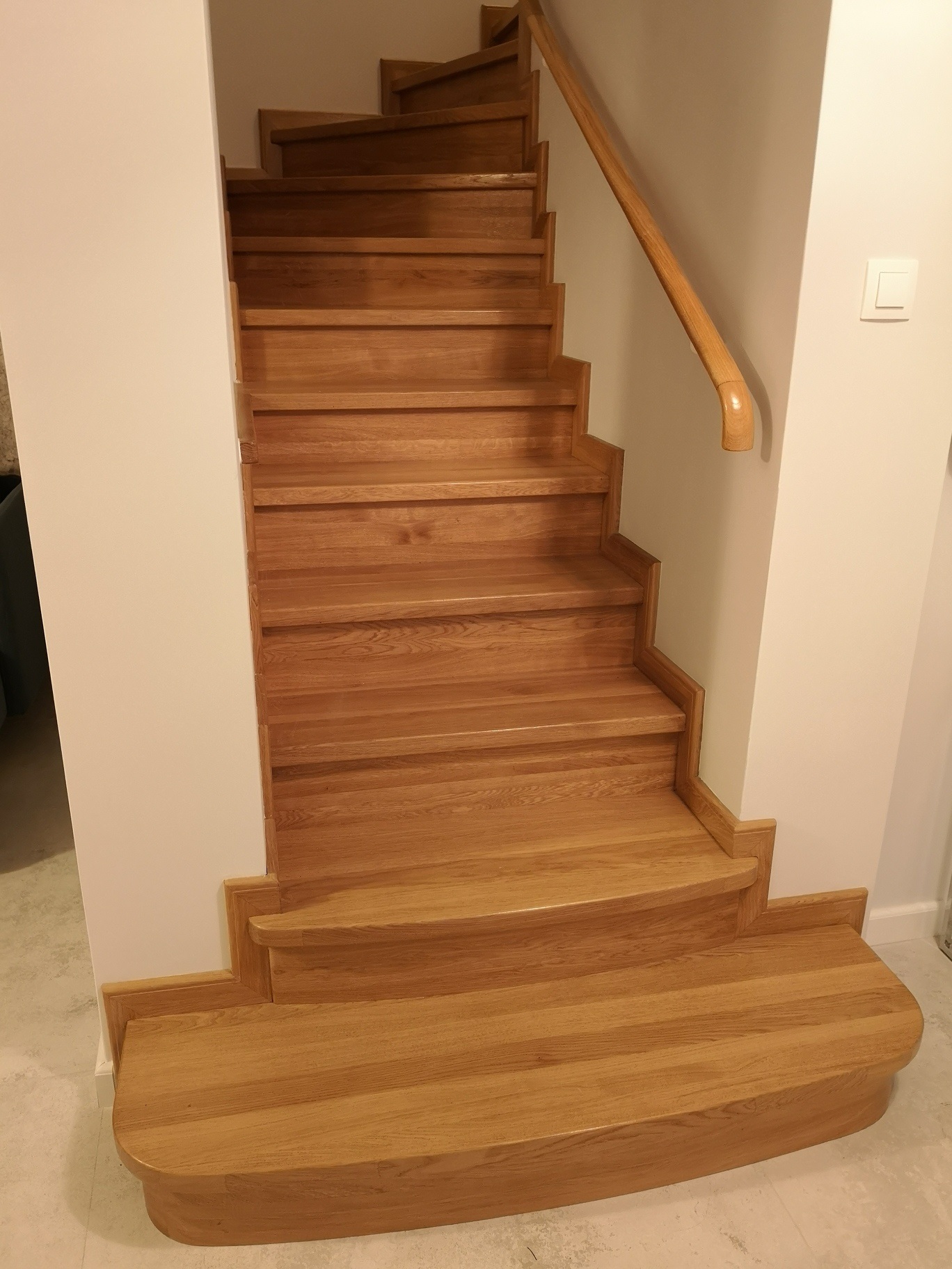 Drewniane schody z jasnego drewna z zaokrąglonym pierwszym stopniem i drewnianą poręczą przy białej ścianie.