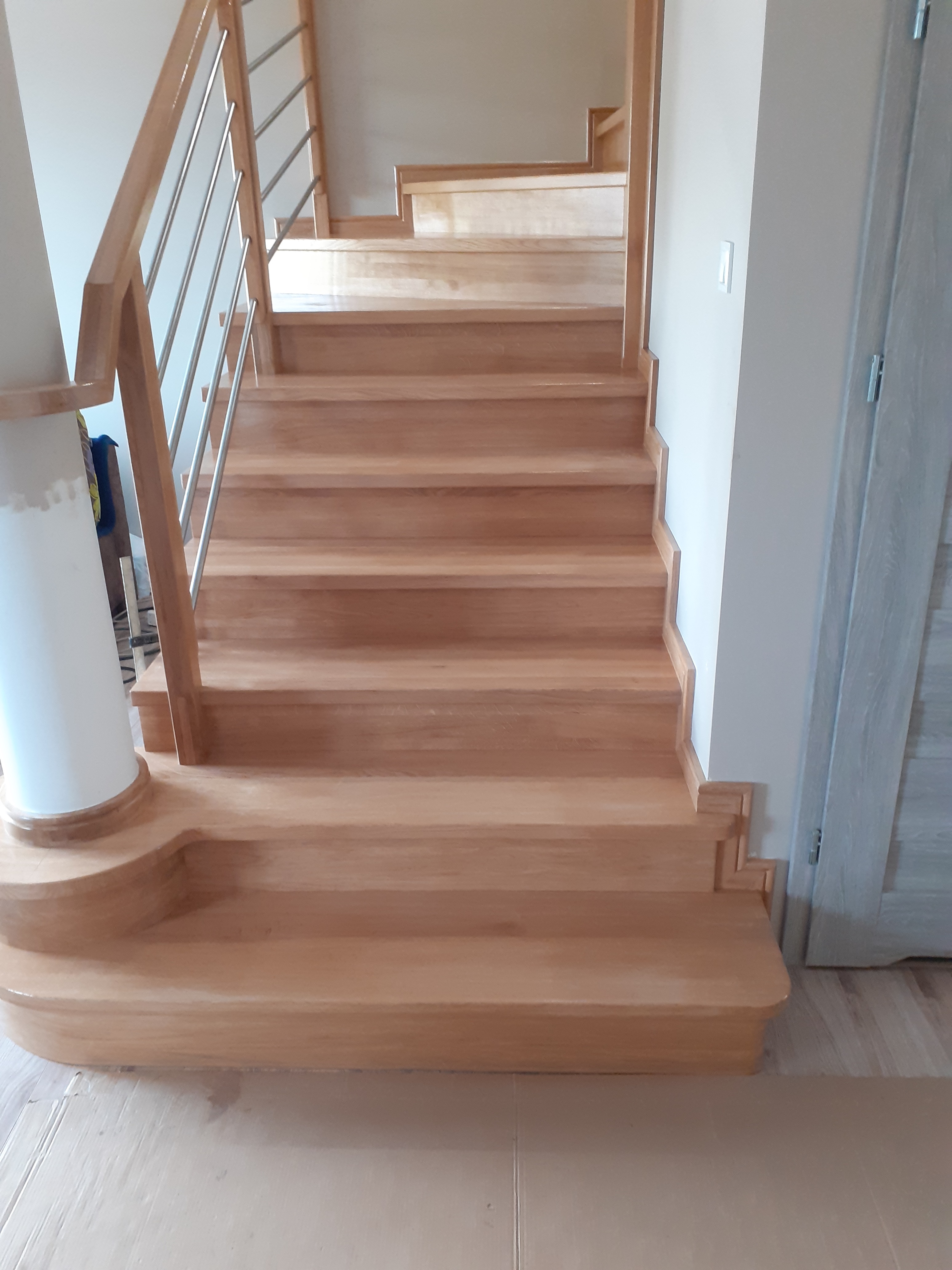 Jasne, drewniane schody z metalową balustradą, widok z dołu na górę, z zaokrąglonym pierwszym stopniem i eleganckim wykończeniem.