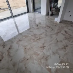 Świeżo położona, błyszcząca podłoga z płytek imitujących marmur w jasnych odcieniach beżu i brązu, odbijająca światło z okien balkonowych, z widocznymi workami z zaprawą w tle.