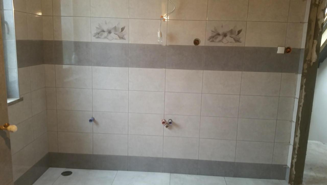 Ściana łazienki wyłożona jasnymi płytkami z szarym pasem dekoracyjnym i motywem kwiatowym, widoczne wystające rury instalacyjne w kolorze niebieskim i czerwonym, oraz surowy stan wykończenia przy...