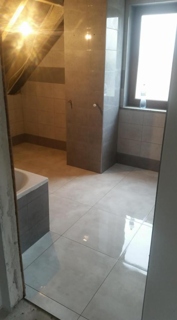 Wykończona łazienka na poddaszu z szarymi płytkami, widoczne skosy dachu i okno z butelką wody na parapecie.