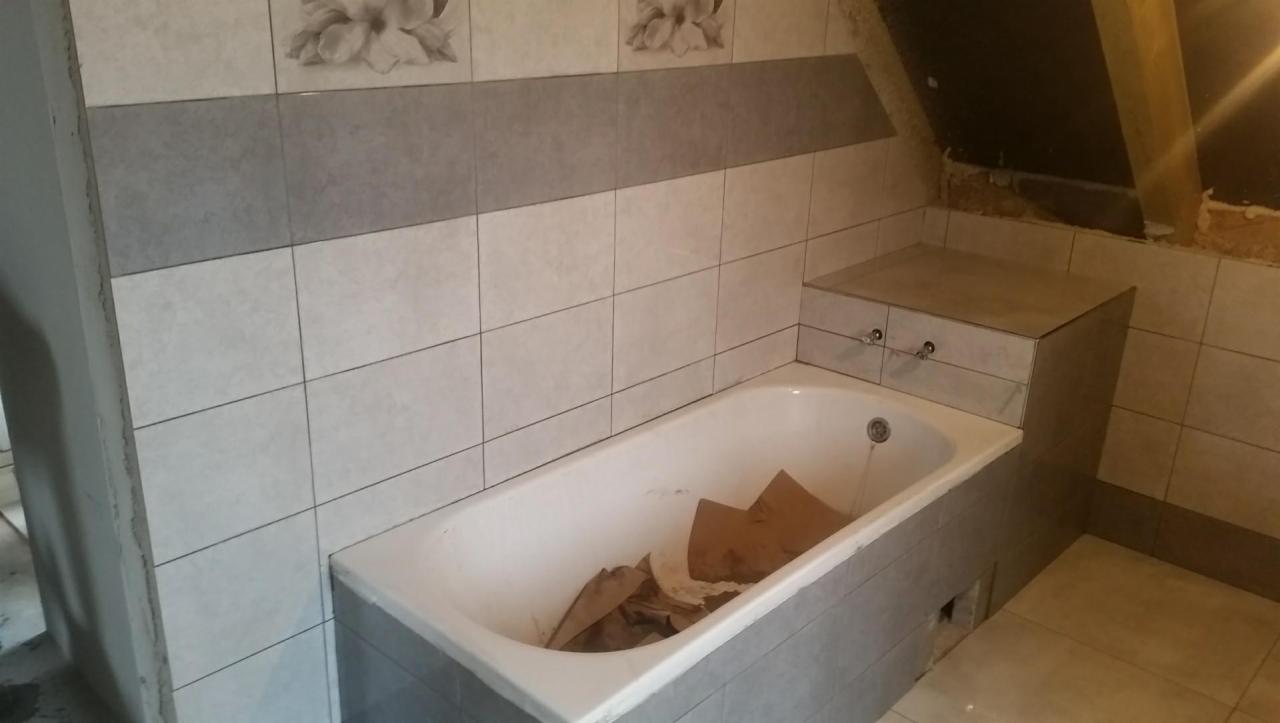 Łazienka w trakcie remontu na poddaszu: białe i szare płytki z motywem kwiatowym, zabudowana wanna z kartonami w środku, widoczne elementy konstrukcyjne dachu.