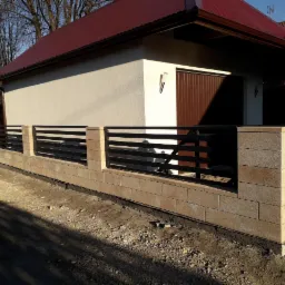 Nowoczesne ogrodzenie z poziomych, czarnych metalowych przęseł osadzonych na słupkach z beżowych bloczków betonowych, na tle budynku z brązowym dachem i bramą garażową.