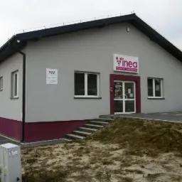 Szary budynek Centrum Medycznego Inea z bordową podmurówką, białymi oknami i schodami wejściowymi, widziany z zewnątrz w pochmurny dzień.