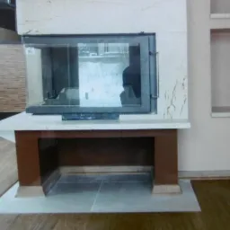 Nowoczesny kominek z marmurowym obramowaniem i brązową podstawą, wkomponowany we wnękę ściany, z widocznym drewnianym parkietem na podłodze.