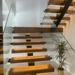 Nowoczesne schody ażurowe z drewnianymi stopniami i szklaną balustradą, oświetlone od dołu, obok doniczka z rośliną, minimalistyczne wnętrze.