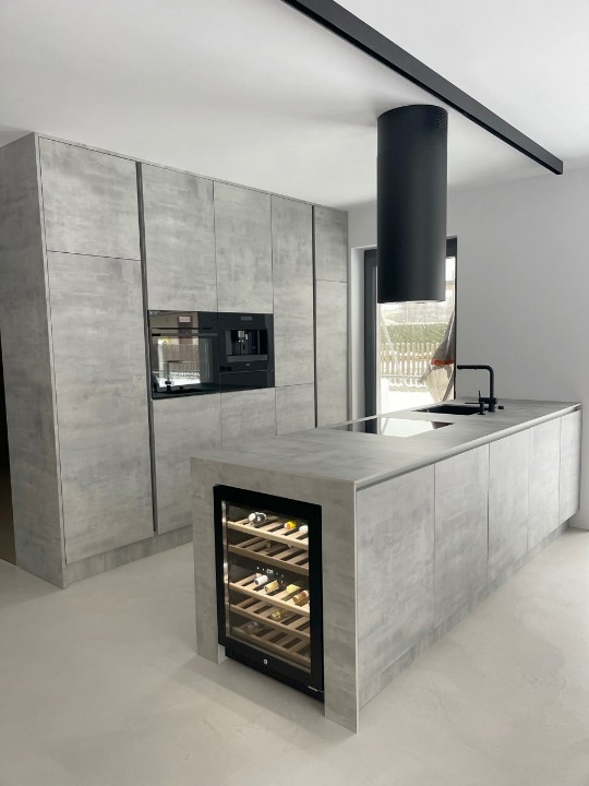 Nowoczesna kuchnia z szarymi, betonowymi frontami szafek, czarnym okapem cylindrycznym i wbudowaną chłodziarką do wina w wyspie.