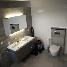 Nowoczesna łazienka z podwójną umywalką, podświetlanym lustrem, toaletą w zabudowie i szarą glazurą imitującą kamień.