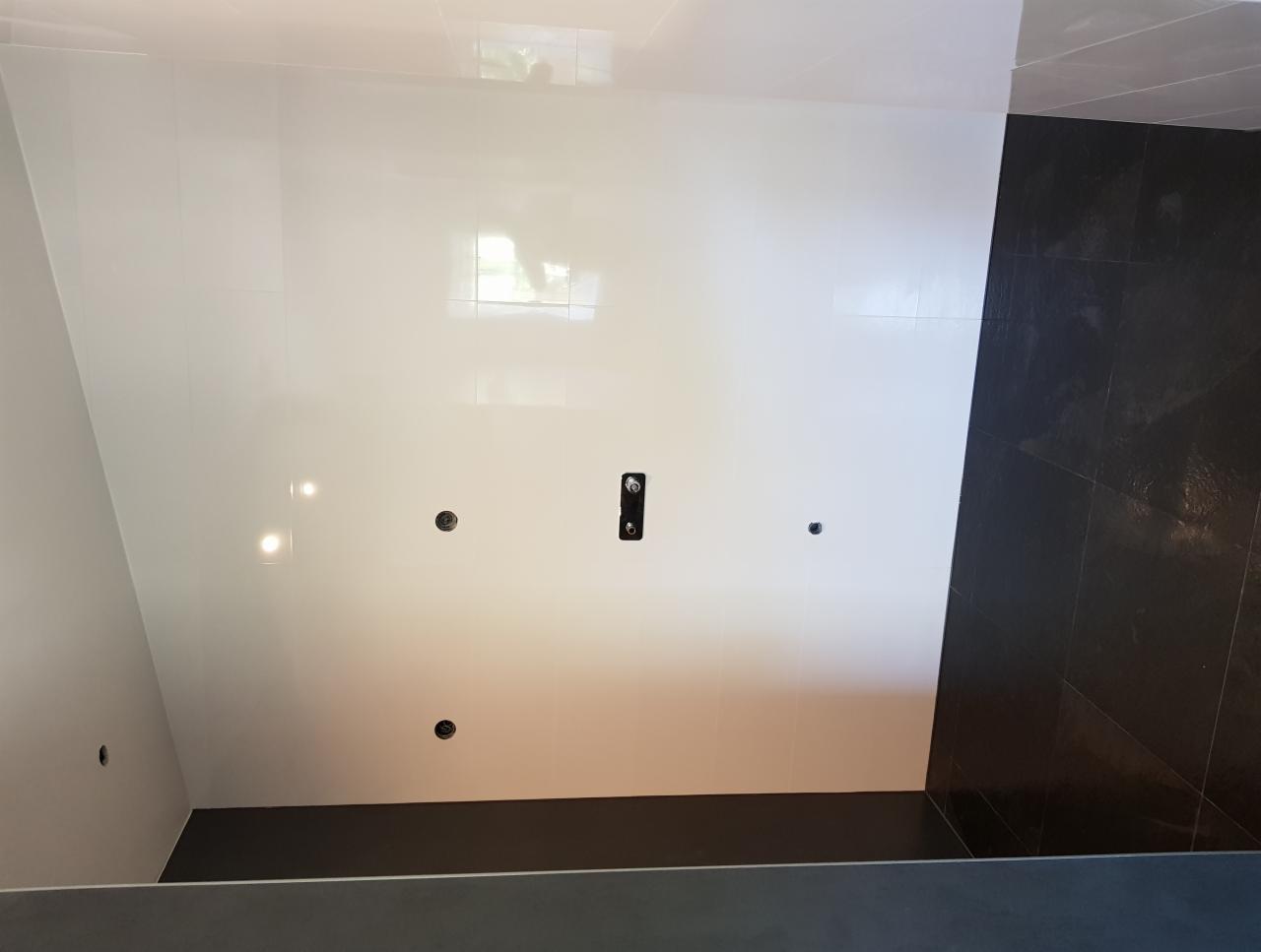 Ściana łazienki wyłożona białymi, błyszczącymi płytkami z widocznymi otworami na armaturę i czarną, matową strefą prysznicową z ciemnymi płytkami podłogowymi.