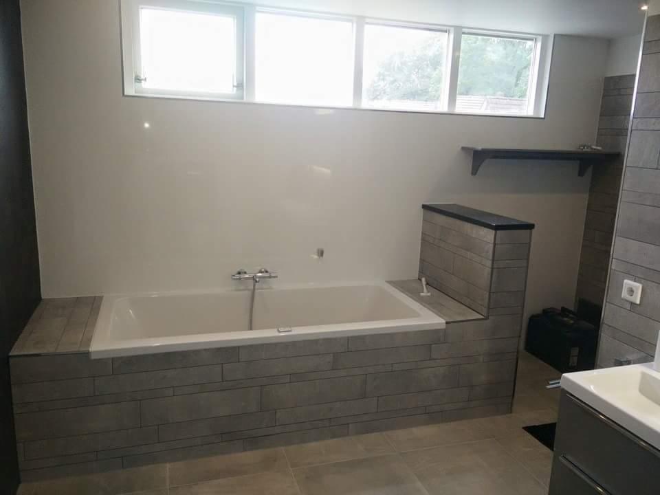 Wykończona łazienka z wanną obudowaną szarą, poziomą mozaiką, białą ścianą z oknem i szarą podłogą z płytek.