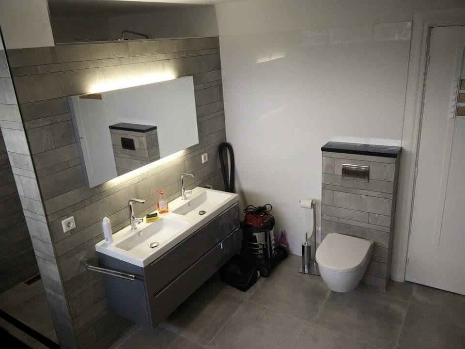 Nowoczesna łazienka z podwójną umywalką, podświetlanym lustrem, toaletą w zabudowie i szarą glazurą imitującą kamień.