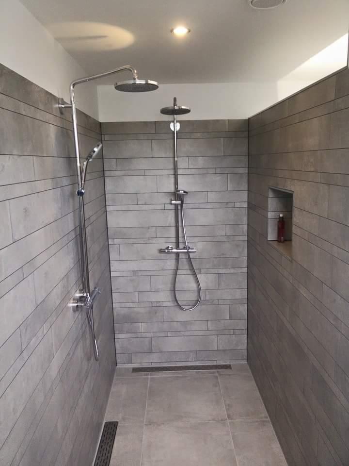 Nowoczesna kabina prysznicowa z szarymi płytkami imitującymi kamień, dwoma deszczownicami i odpływem liniowym w podłodze.