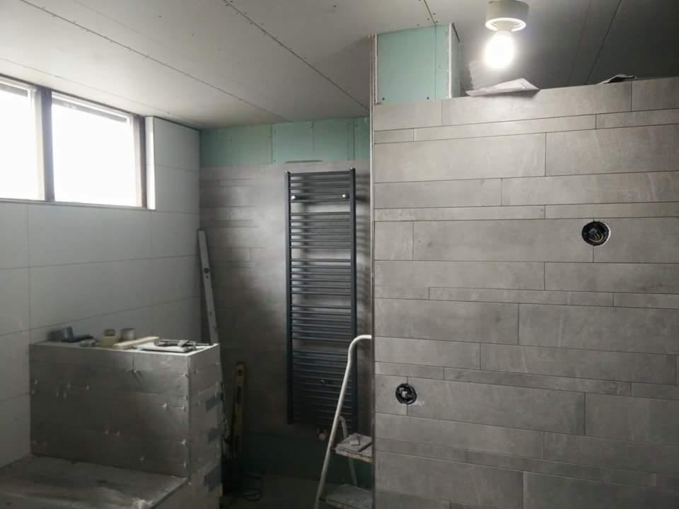 Łazienka w trakcie remontu: ściany wyłożone szarymi, podłużnymi płytkami, grzejnik drabinkowy, zabudowa w folii aluminiowej, surowy sufit z widocznymi łączeniami płyt gipsowych, drabina i poziomica.