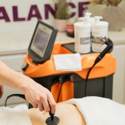 FizjoBalance - Zabieg z użyciem aparatu do fizykoterapii na brzuchu pacjenta, widoczna dłoń terapeuty i pomarańczowy sprzęt w gabinecie.