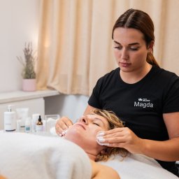 FizjoBalance - Fizjoterapeutka Magda oczyszcza twarz klientki w gabinecie, używając wacików. Widoczne kosmetyki, neutralne tło i logo firmy na koszulce.