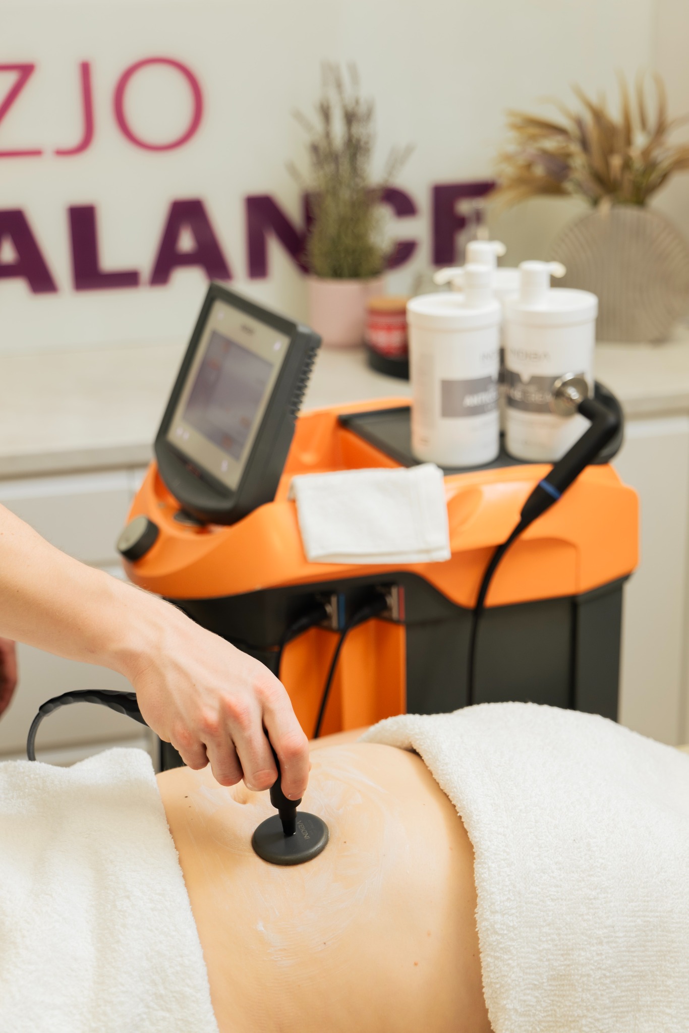 Zabieg z użyciem aparatu do fizykoterapii na brzuchu pacjenta, widoczna dłoń terapeuty i pomarańczowy sprzęt w gabinecie.