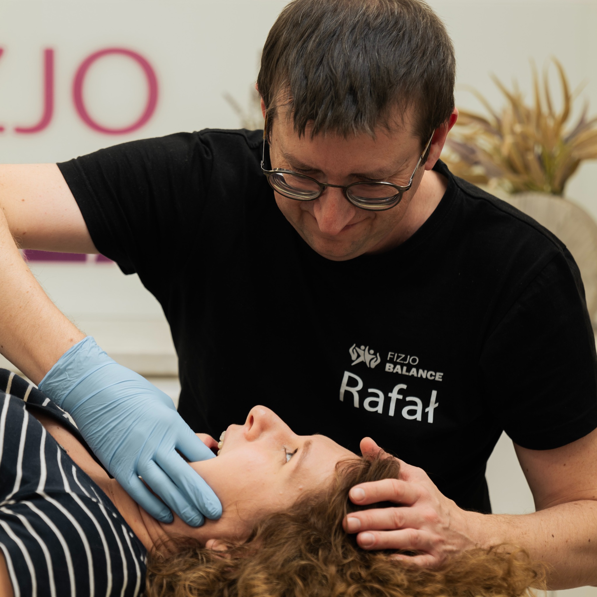 Mężczyzna w czarnej koszulce z logo 'Fizjo Balance Rafał' w niebieskich rękawiczkach wykonuje masaż szyi pacjentki leżącej na plecach. Ujęcie z bliska.