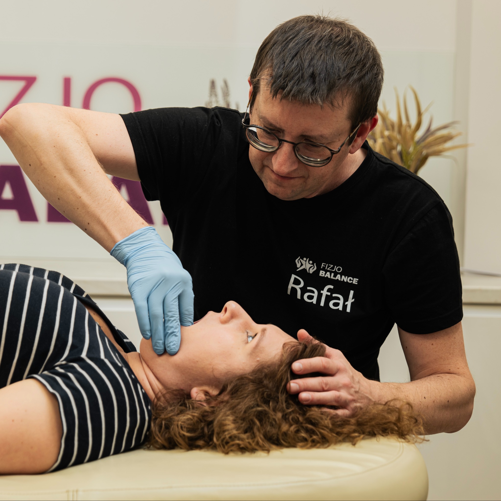 Sesja manualna: terapeuta w okularach w niebieskich rękawiczkach delikatnie masuje szyję pacjentki leżącej na leżance. Widoczne logo 'Fizjo Balance Rafał'.