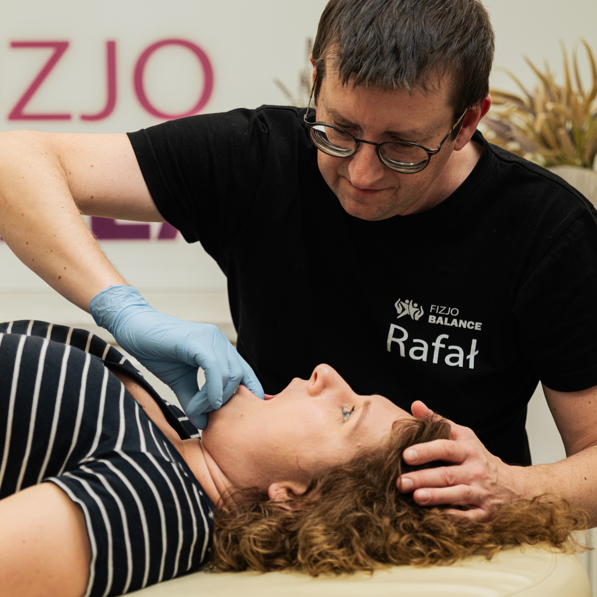 Terapia manualna: terapeuta w rękawiczkach bada jamę ustną pacjentki leżącej na kozetce. Widoczne logo 'Fizjo Balance Rafał' na koszulce terapeuty.