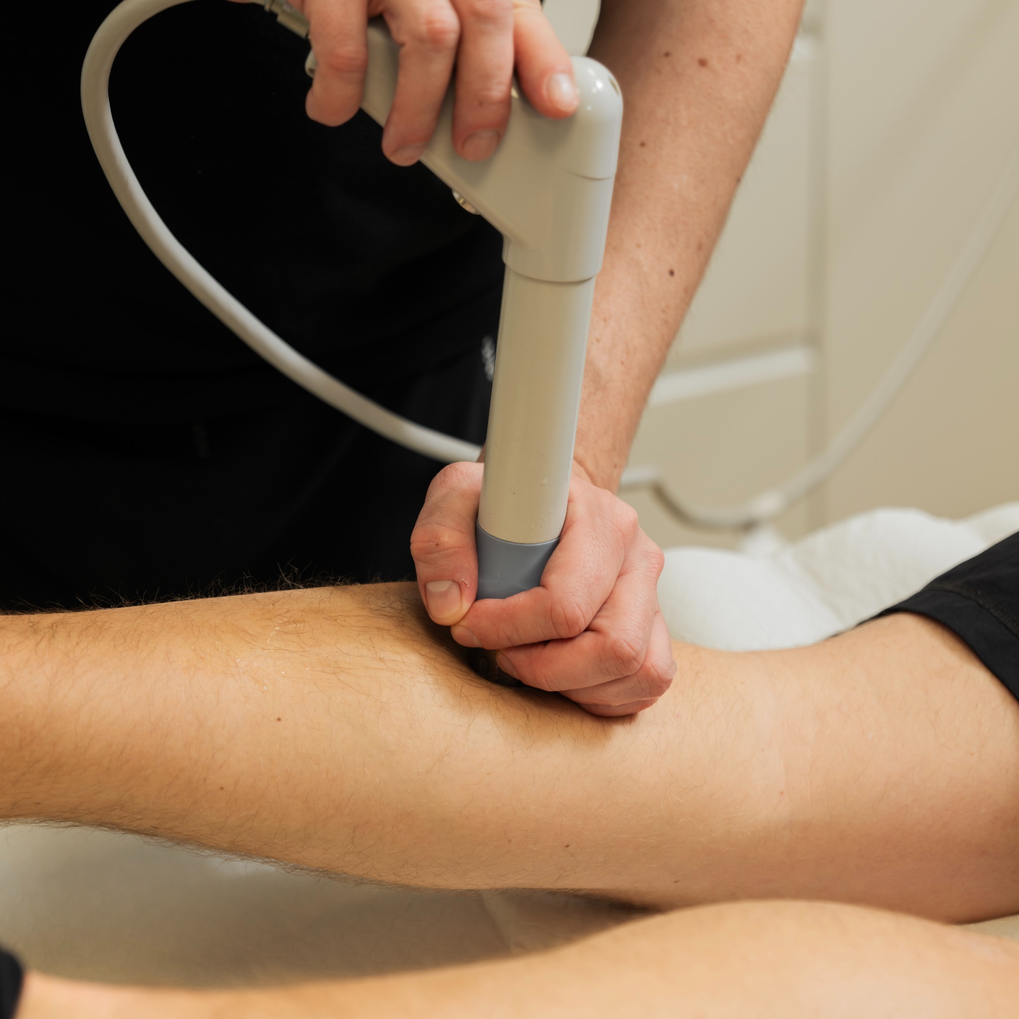 Zabieg ultradźwiękowy na łydce pacjenta, wykonywany przez osobę w czarnym ubraniu. Widoczny aplikator i kabel urządzenia, skóra z delikatnym owłosieniem.