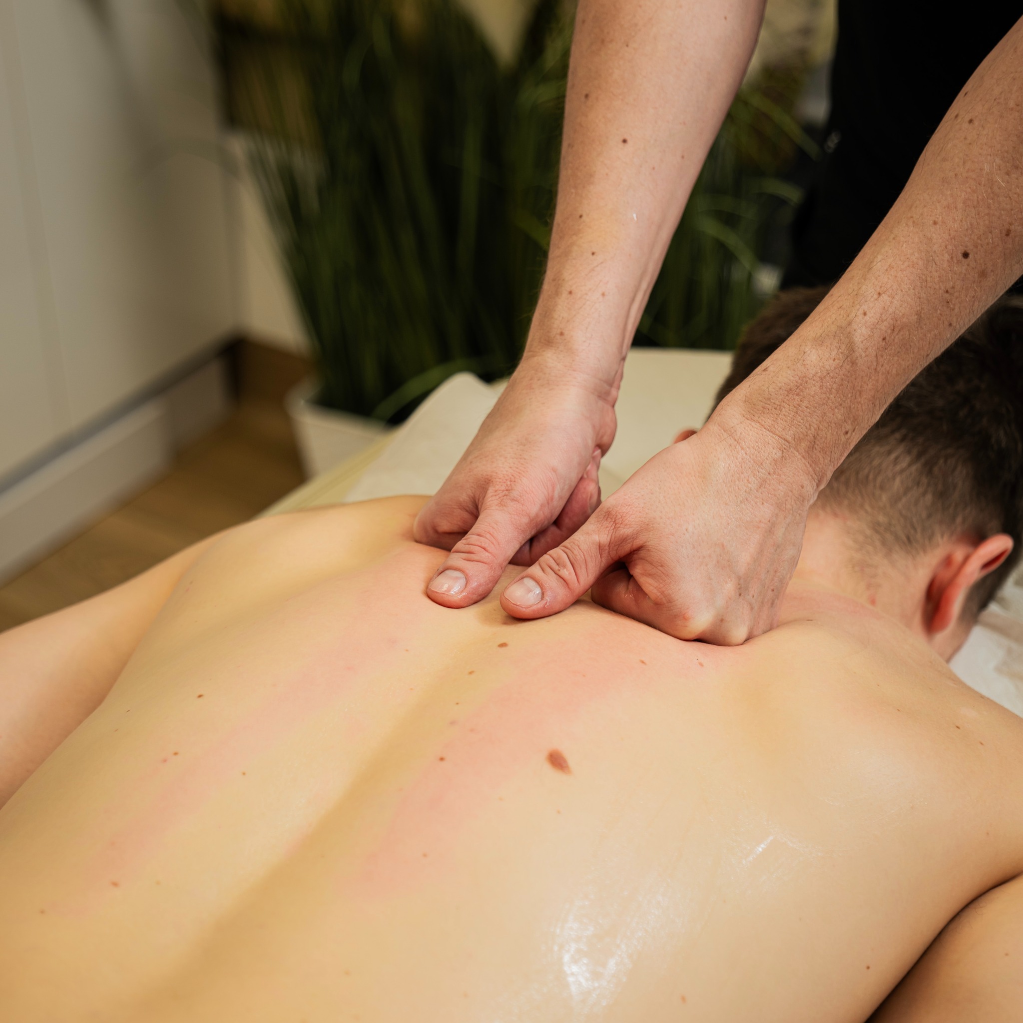 Masaż pleców pacjenta wykonywany dłońmi, widoczne zaczerwienienia skóry po zabiegu, w tle zielona roślina doniczkowa i fragment białego prześcieradła.