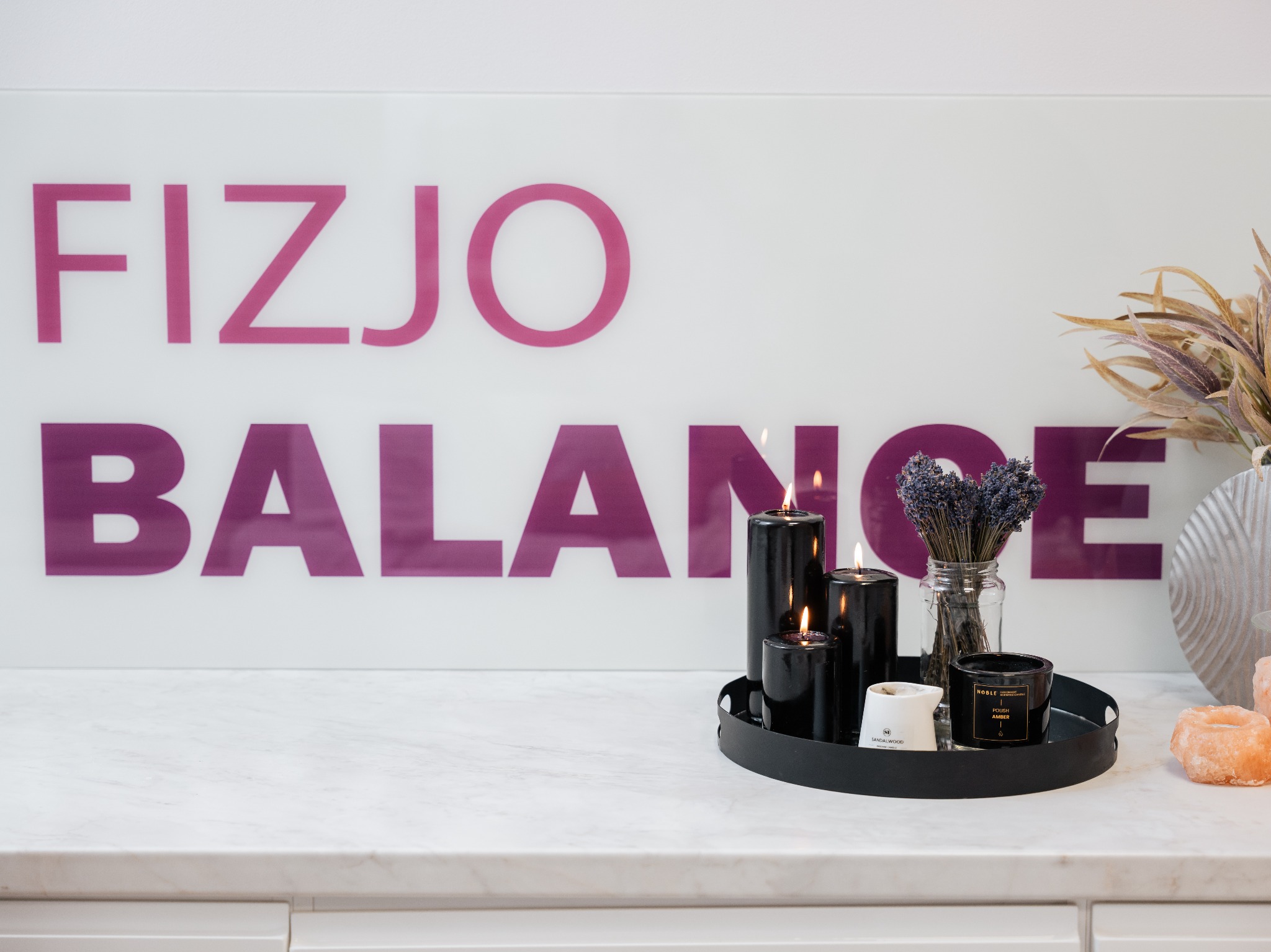 Stylowe wnętrze gabinetu z napisem 'Fizjo Balance' w tle. Na marmurowym blacie czarne świece, lawenda w słoiku i dekoracje w odcieniach bieli i różu.