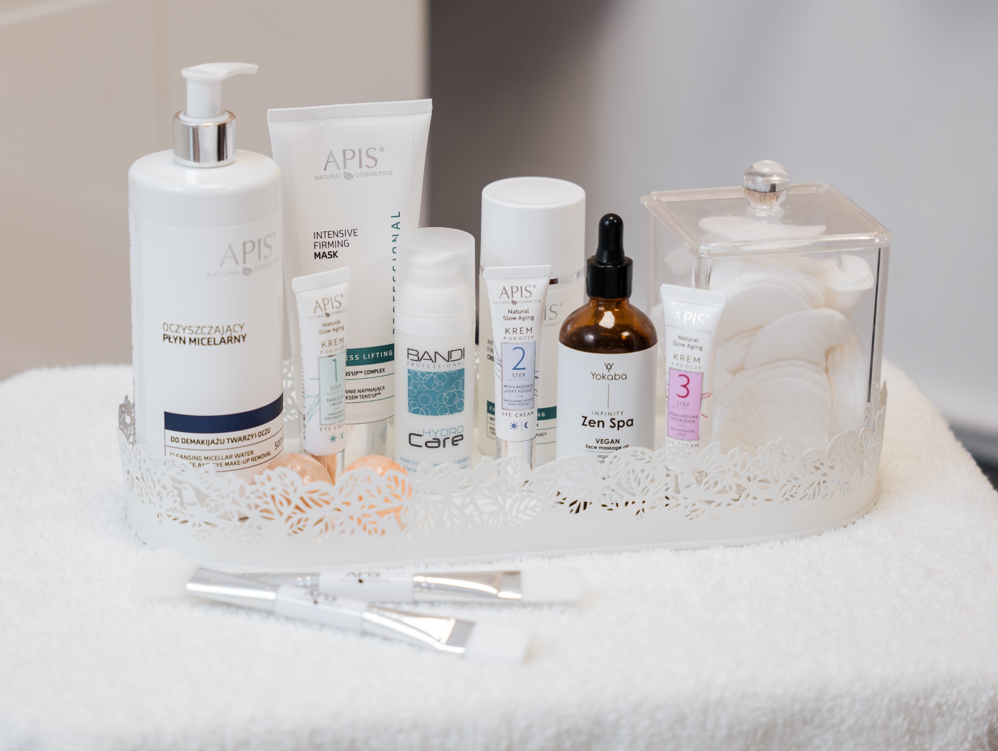 Zestaw kosmetyków do pielęgnacji twarzy różnych marek, w tym APIS i Bandi, na białej, ażurowej tacy. Widoczne płyny micelarne, kremy pod oczy i olejki.