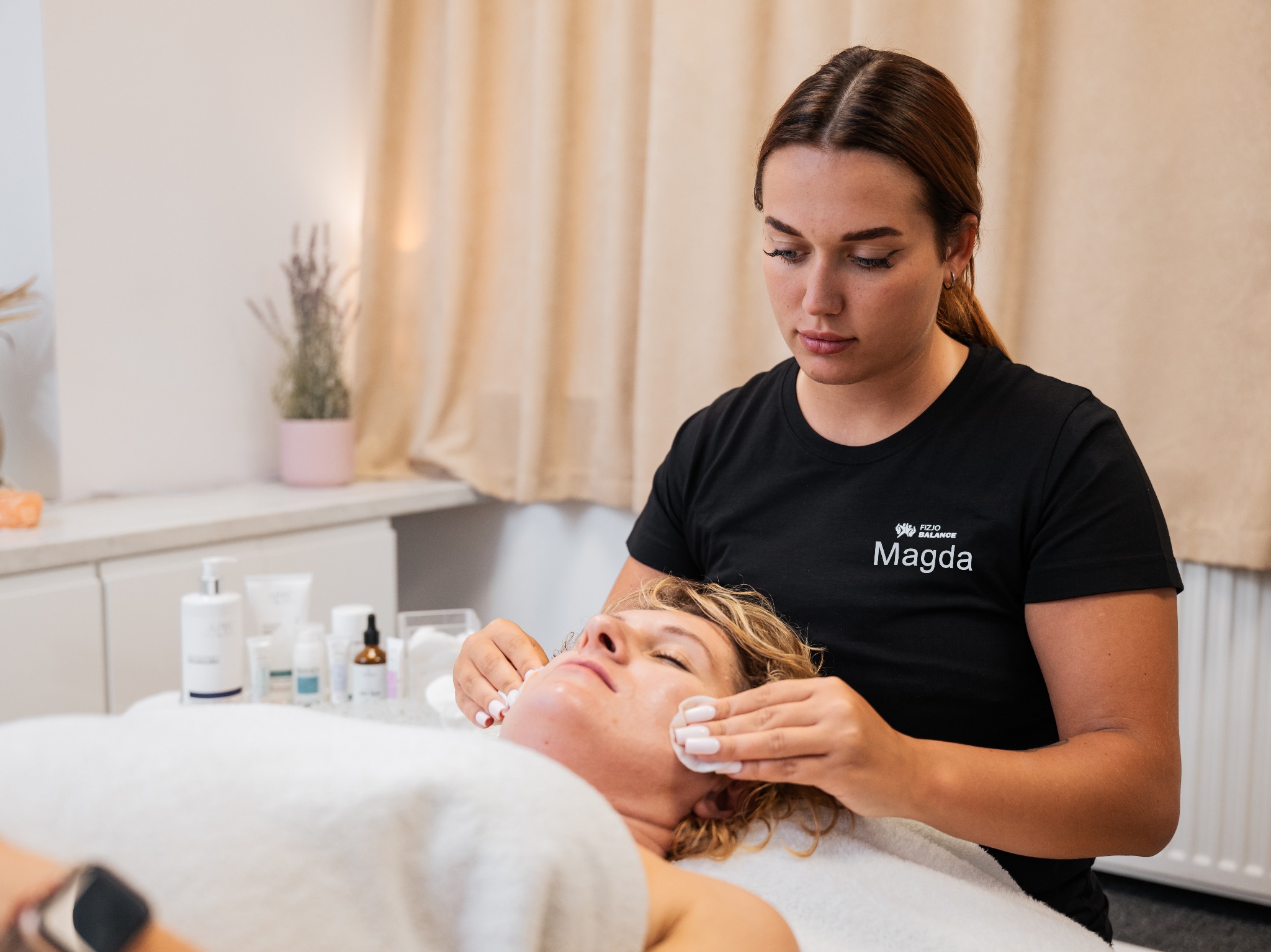 Fizjoterapeutka Magda oczyszcza twarz klientki w gabinecie, używając wacików. Widoczne kosmetyki, neutralne tło i logo firmy na koszulce.
