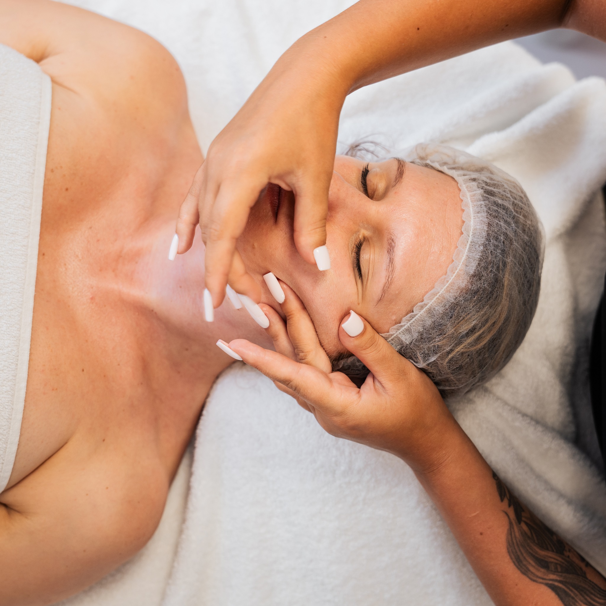 Kobieta w czepku poddawana masażowi twarzy. Dłonie z długimi, białymi paznokciami delikatnie uciskają skórę. Białe ręczniki w tle. Zabieg upiększający.