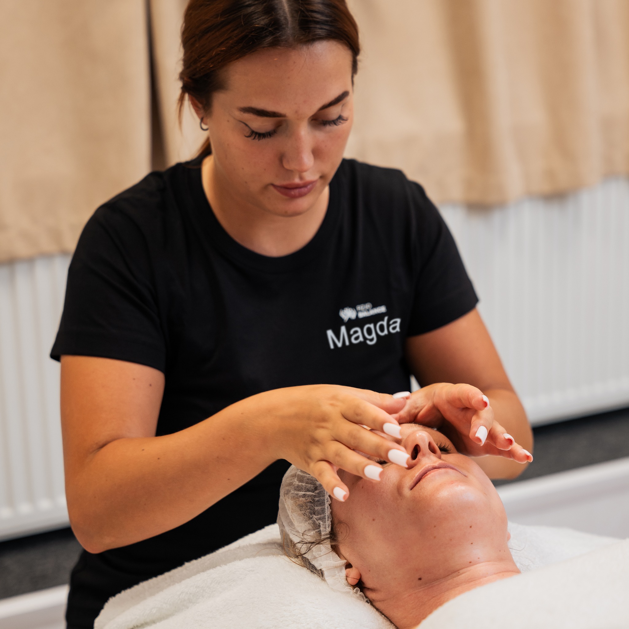 Osoba wykonuje masaż twarzy klientce leżącej na leżance. Kobieta w czarnej koszulce z logo firmy delikatnie masuje skórę twarzy pacjentki.