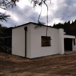 Nowoczesny dom jednorodzinny z białą elewacją i czarnymi akcentami, widoczny z zewnątrz, z fragmentem iglastego drzewa w kadrze.