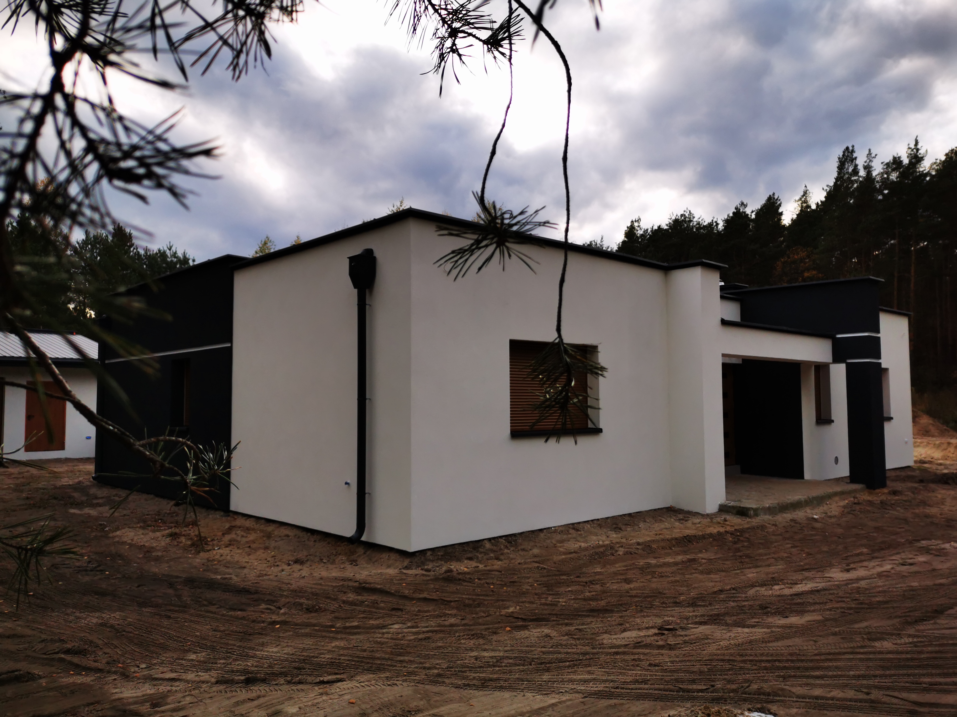 Nowoczesny dom jednorodzinny z białą elewacją i czarnymi akcentami, widoczny z zewnątrz, z fragmentem iglastego drzewa w kadrze.