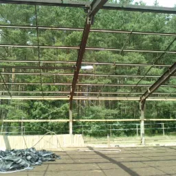 Konstrukcja stalowa hali w trakcie budowy, widoczne elementy drewniane i sterta czarnych worków na ziemi, w tle las.