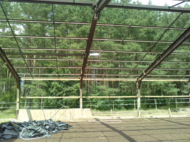 Konstrukcja stalowa hali w trakcie budowy, widoczne elementy drewniane i sterta czarnych worków na ziemi, w tle las.