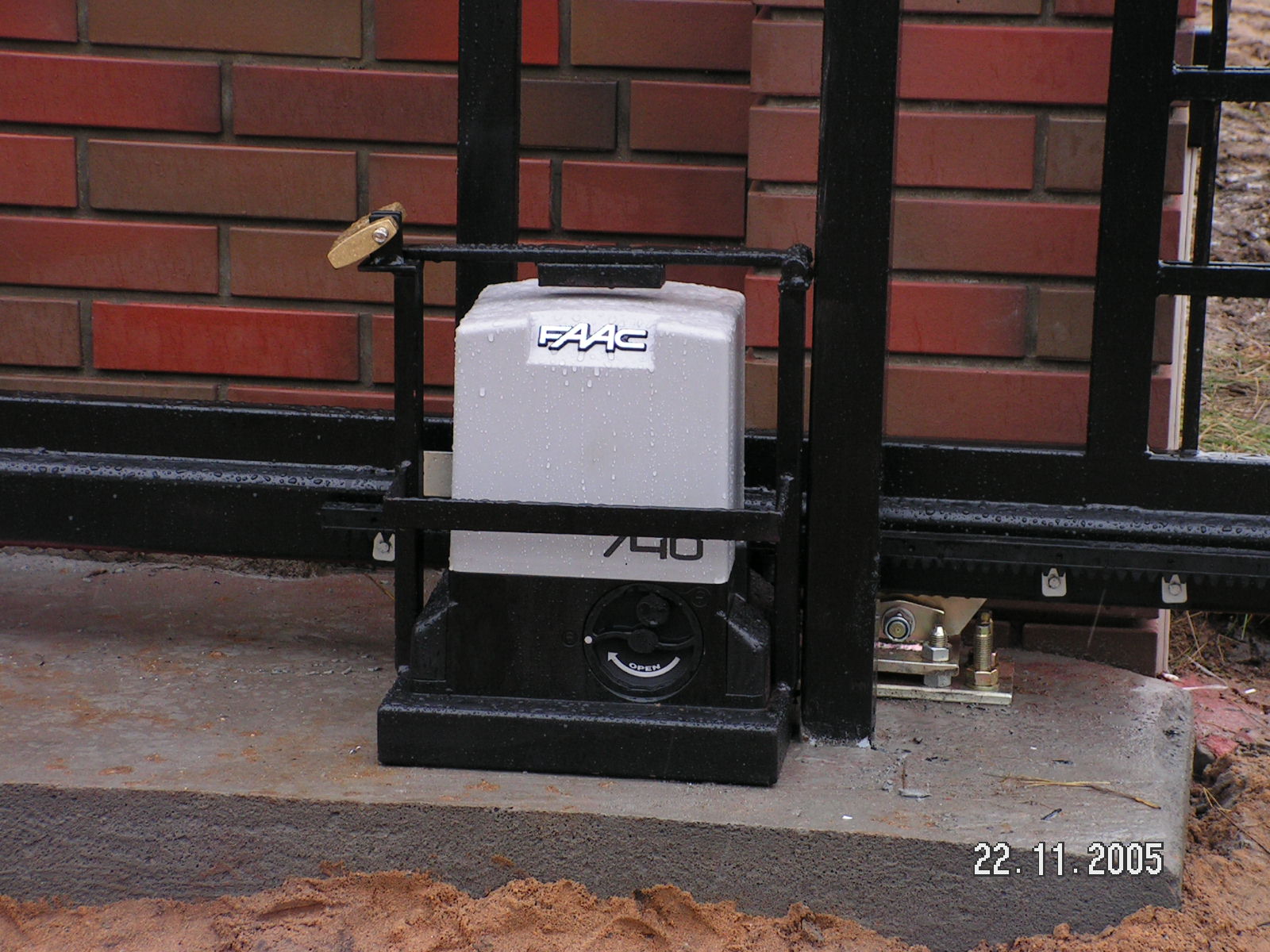 Automat FAAC do bramy przesuwnej, zabezpieczony kłódką, zamontowany na betonowym fundamencie, z widoczną zębatką i ceglanym murem w tle, wykonany w pochmurny dzień.