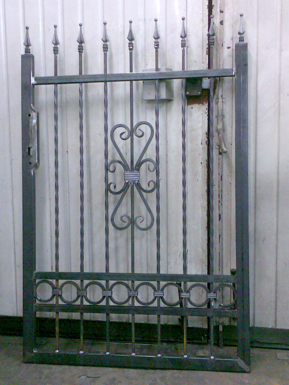 Surowa, metalowa furtka z dekoracyjnymi elementami w kształcie serc i okręgów, widoczne ślady spawania i niedoskonałości wykończenia, ustawiona na tle blaszanej ściany.