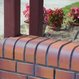 Detail of a brick fence base with dark brown metal posts, showing a welded connection, with potted pink flowers in the background.