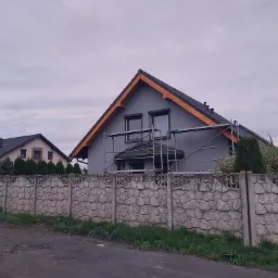 Dom jednorodzinny w trakcie malowania elewacji na szaro, widoczne rusztowania i fragment ozdobnego betonowego ogrodzenia.