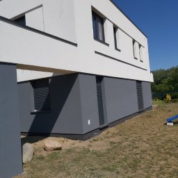 MAX-BUD - Nowoczesny dom jednorodzinny z białą elewacją i szarą podmurówką w Rogoźnie, widok z boku, na trawniku leżą kamienie, w tle zieleń drzew.