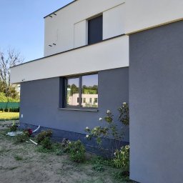 MAX-BUD - Nowoczesny dom z szarą elewacją, widok z boku, fragment ogrodu z młodymi roślinami i sprzętem ogrodniczym.